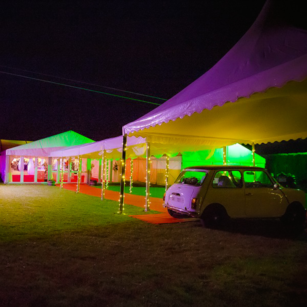 A wedding marquee entrance tunnel photographed at night, with coloured lights and a classic mini car parked by the entrance tunnel